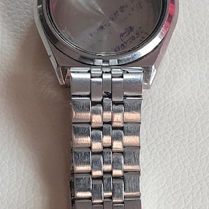 May include: A silver-toned metal watch case with an attached multi-link metal bracelet. The watch face is missing, revealing an interior surface with handwritten text that includes the date 25.02.2015 and a small signature in blue ink.