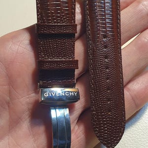 May include: A brown watch strap with a textured lizard-skin pattern. The strap features a silver-toned metal deployment clasp with the brand name GIVENCHY engraved on the surface. The two-piece strap includes a buckle side and a perforated tail end.