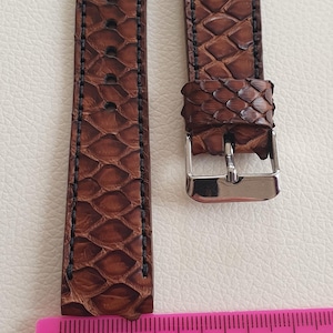 May include: A brown watch strap with a textured scale pattern and black stitching, featuring a silver-toned metal buckle. A pink plastic ruler is positioned at the base, showing the strap length measures approximately 5 inches in total.