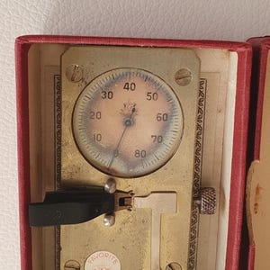 May include: A vintage brass Favorite Swiss Made watchmaker tool housed in a red box. The device features a circular gauge with a needle pointing near zero, a black lever, a silver T-shaped component, and a small knurled knob on the side.