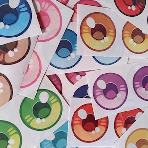 Puede incluir: Una colección de pegatinas de ojos coloridos en varios tonos, incluyendo azul, rosa, marrón, verde, naranja, rojo y morado. Cada pegatina presenta un diseño detallado de iris y pupila, impreso en un material blanco texturizado. Las pegatinas están dispersas.