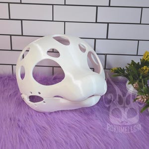 May include: A white, open-faced, 3D-printed mask with large, circular eye and mouth openings. The mask has a smooth, rounded design with several smaller holes across the top. The mask is designed to be worn as part of a costume.