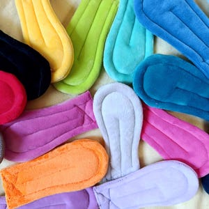 May include: A collection of colorful, plush, reusable sanitary pads in various shades including black, yellow, green, blue, pink, and orange. The pads have a contoured shape with a soft, absorbent surface, designed for comfort and protection.