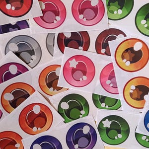 Puede incluir: Una colección de pegatinas de ojos coloridos en tonos de rojo, rosa, naranja, verde, azul, morado y marrón. Cada pegatina presenta un diseño detallado de iris y pupila, algunas con acentos de estrella. Las pegatinas están sobre un fondo blanco.
