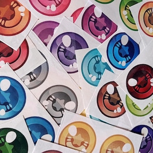 Puede incluir: Una colección de pegatinas de ojos de estilo caricatura y coloridas. Las pegatinas presentan una variedad de colores, incluyendo rojo, azul, morado, verde y naranja. Cada ojo tiene un reflejo blanco y una pupila en forma de estrella. Las pegatinas están sobre un fondo blanco.