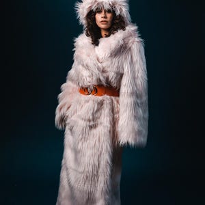 May include: A long, pink faux fur coat with a large hood. The coat has a brown belt with a gold buckle.