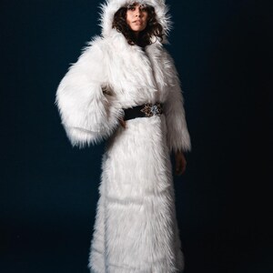 May include: A white faux fur coat with a hood. The coat is long and has a belt around the waist.