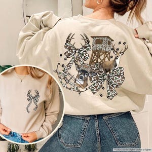 May include: Sand-colored sweatshirt featuring a deer-themed design. The back of the sweatshirt showcases a deer, bow, and hunting blind with camouflage bows. A smaller version of the design is on the front. The text "Sand" is in the upper left corner.
