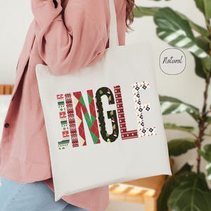 May include: A white tote bag with the word "JINGLE" printed in a festive, patterned design. The letters are decorated with red, green, and black patterns, creating a holiday theme. The bag is carried by a person wearing a pink shirt.