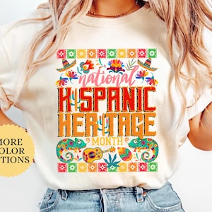 May include: Ivory-colored t-shirt with a colorful graphic celebrating National Hispanic Heritage Month. The design includes the text "National Hispanic Heritage Month" with decorative elements like sombreros, flowers, and chameleons. The shirt is a casual wear item.
