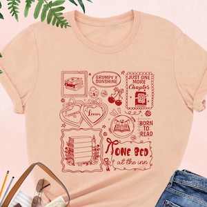 May include: Heather peach t-shirt with a red graphic design featuring book-themed illustrations and text. The design includes phrases like "Grumpy x Sunshine," "Just One More Chapter," and "Born to Read." The shirt is paired with blue jeans and a brown handbag.