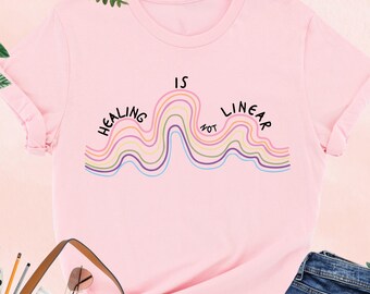 May include: Pink t-shirt with the text "Healing is not linear" in a wavy design. The shirt is displayed with a brown tote bag, sunglasses, a camera, and blue jeans. Green leaves and foliage surround the items.