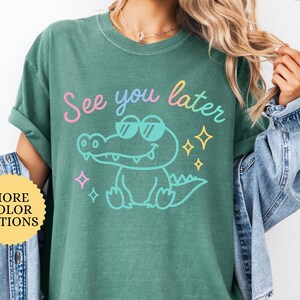 Puede incluir: Una camiseta verde claro con la frase "See you later" y un cocodrilo de dibujos animados con gafas de sol. La camiseta tambi&eacute;n tiene un bot&oacute;n que dice "M&aacute;s opciones de color".