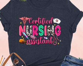 May include: Heather navy t-shirt with the words "Certified Nursing Assistant" in colorful, playful lettering. The design includes medical symbols and a nurse's cap. A pair of blue jeans and a brown tote bag are also in the image.