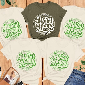 May include: Four t-shirts with the text 'All Day Every Day Mental Health Matters' in green lettering on a white circle. The shirts are laid out on a wooden surface with green plants and a brown leather bag in the background. The shirts are available in natural and heather olive colors.