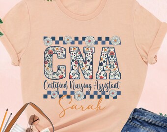 May include: A peach colored t-shirt with a floral design that says "CNA Certified Nursing Assistant" and the name "Sarah" underneath.