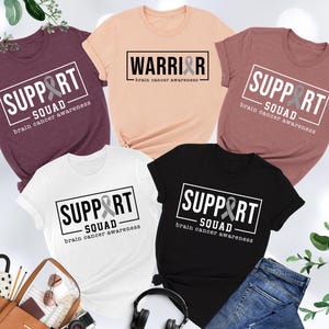 May include: Four t-shirts with different designs and colors. The shirts all feature a gray ribbon and the text "Support Squad" or "Warrior" with the words "brain cancer awareness". The shirts are available in heather peach, heather mauve, heather maroon, white, and black.