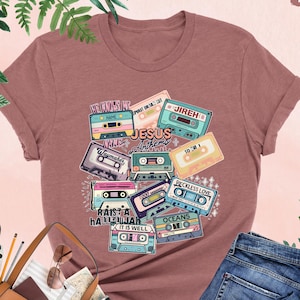 May include: Heather mauve t-shirt featuring a graphic of vintage cassette tapes with the words "Jesus Anthems" and other song titles. The design includes various colors and fonts. The shirt is paired with blue jeans and accessories.