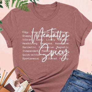 K&ouml;nnte beinhalten: Heather-Mauve-T-Shirt mit dem Schriftzug "Mentally Spicy" in wei&szlig;er Schrift. Das Shirt zeigt eine Liste beschreibender W&ouml;rter. Eine braune Handtasche, Sonnenbrille und Jeans sind im Bild.