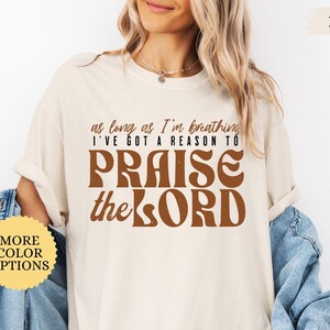 May include: A white t-shirt with the text "as long as I'm breathing I've got a reason to PRAISE the LORD" in brown letters. The shirt is being modeled by a person.
