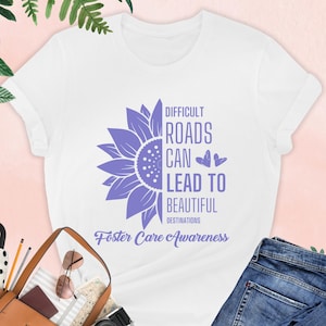 May include: White t-shirt with a purple sunflower graphic and the text "Difficult Roads Can Lead To Beautiful Destinations Foster Care Awareness".