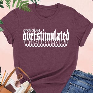 May include: Heather maroon t-shirt with white text that reads "probably overstimulated so please don't talk to me" with a checkered pattern underneath the text.