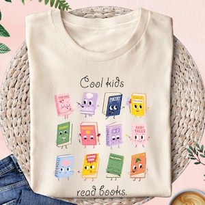 Puede incluir: Camiseta color crema con el texto "Cool kids read books" y personajes de libros de dibujos animados. Los libros son de varios colores y est&aacute;n etiquetados con g&eacute;neros como "Romance", "Poetry" y "Fairy Tales".