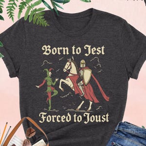 May include: Dark grey heather t-shirt with the text "Born to Jest Forced to Joust" and a graphic of a jester, a knight on a horse, and a shield. The shirt is displayed with a brown tote bag, sunglasses, and blue jeans.