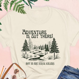 May include: Cream-colored t-shirt with the text "ADVENTURE IS OUT THERE! BUT SO ARE SERIAL KILLERS" in a dark green font. The graphic features a forest scene with a winding river. A pair of blue jeans and a brown purse are also visible.
