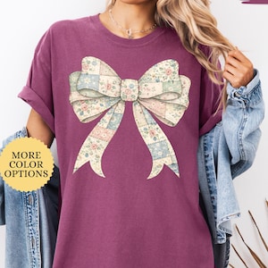 May include: Berry-colored t-shirt featuring a large bow graphic. The bow is designed with a patchwork pattern of floral and pastel squares. The shirt has rolled sleeves and the text "MORE COLOR OPTIONS" is visible.