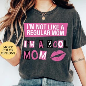 May include: Dark gray t-shirt with the text "I'M NOT LIKE A REGULAR MOM I'M A COOL MOM" in pink and black lettering. A pink lip graphic is below the text. The shirt has a crew neck and short sleeves.