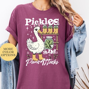 May include: Berry-colored t-shirt with a whimsical graphic featuring a goose wearing a bow and boots, alongside jars of pickles. The text reads "Pickles and Panic Attacks". Additional text indicates more color options.