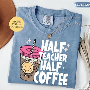 May include: A blue jean Comfort Colors t-shirt with the text "HALF TEACHER HALF COFFEE" in white. The graphic includes a pink-lidded coffee cup with a smiley face and a black and white straw. A gold necklace is draped across the shirt.
