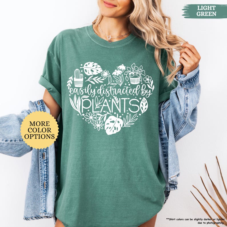 Pu&ograve; includere: Una maglietta verde chiaro con un design a forma di cuore bianco con il testo "easily distracted by PLANTS" e una variet&agrave; di illustrazioni di piante.