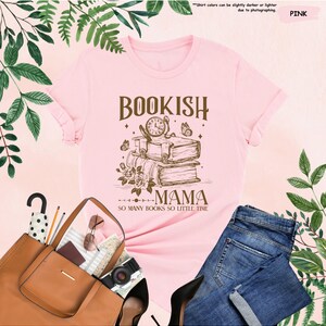 May include: Pink t-shirt with a graphic design featuring the words "BOOKISH MAMA" above a stack of books, a clock, and butterflies. The text "SO MANY BOOKS SO LITTLE TIME" is below the graphic. The shirt is displayed with a brown tote bag and blue jeans.