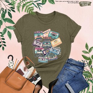 May include: Olive green t-shirt featuring a design of retro cassette tapes with religious phrases like "Jesus Anthems" and "Reckless Love." The shirt is paired with blue jeans, a brown tote bag, and black heels.