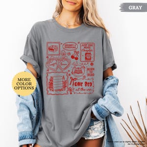 Puede incluir: Camiseta gris con ilustraciones gr&aacute;ficas rojas de libros, corazones y texto. El texto incluye "Grumpy x Sunshine", "Just One More Chapter", "Born to Read" y "One Bed at the Inn". La camiseta tiene mangas enrolladas.
