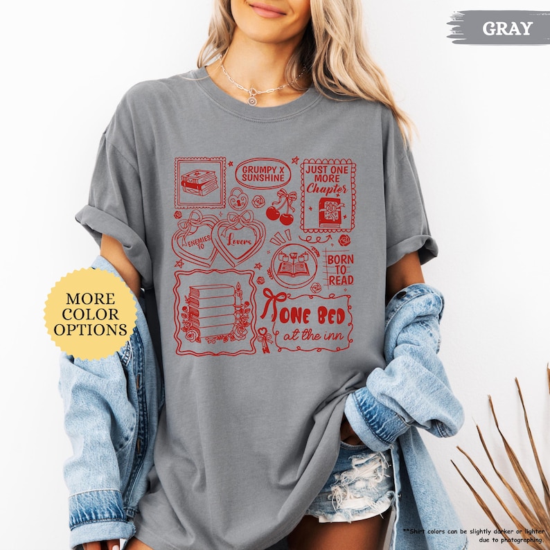 Puede incluir: Camiseta gris con ilustraciones gr&aacute;ficas rojas de libros, corazones y texto. El texto incluye "Grumpy x Sunshine", "Just One More Chapter", "Born to Read" y "One Bed at the Inn". La camiseta tiene mangas enrolladas.