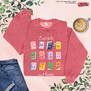 Puede incluir: Sudadera de color rojo escarlata jaspeado con el texto "Cool kids read books". La sudadera presenta un gr&aacute;fico de libros de dibujos animados con caras. Una taza de caf&eacute; y unos vaqueros azules tambi&eacute;n est&aacute;n en la imagen.