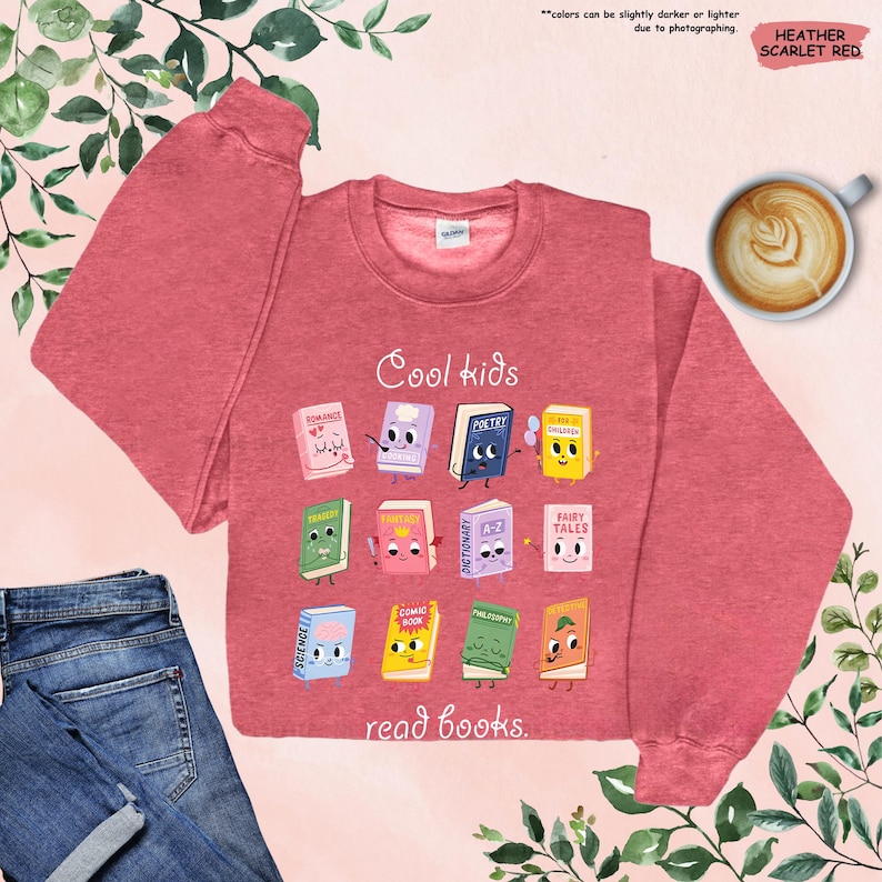 Puede incluir: Sudadera de color rojo escarlata jaspeado con el texto "Cool kids read books". La sudadera presenta un gr&aacute;fico de libros de dibujos animados con caras. Una taza de caf&eacute; y unos vaqueros azules tambi&eacute;n est&aacute;n en la imagen.