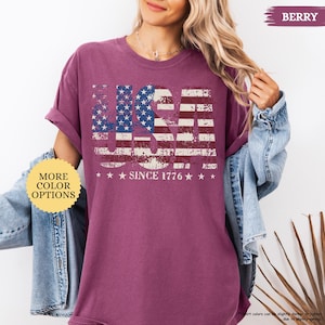 May include: Berry-colored t-shirt with a distressed USA flag design. The design features the word "USA" in a patriotic font, with the American flag incorporated into the letters. Below the flag design, the text reads "SINCE 1776".
