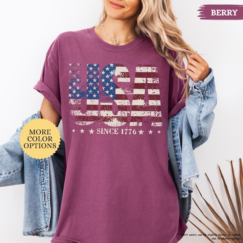 May include: Berry-colored t-shirt with a distressed USA flag design. The design features the word "USA" in a patriotic font, with the American flag incorporated into the letters. Below the flag design, the text reads "SINCE 1776".