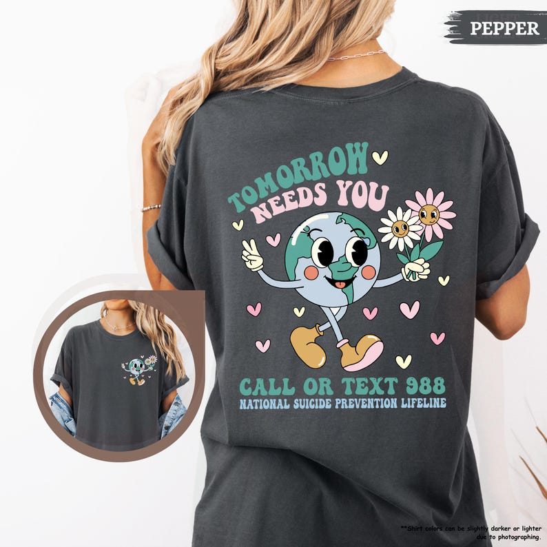 May include: Dark grey t-shirt featuring a cartoon earth character with flowers and the text "TOMORROW NEEDS YOU." The shirt also includes the text "CALL OR TEXT 988 NATIONAL SUICIDE PREVENTION LIFELINE."