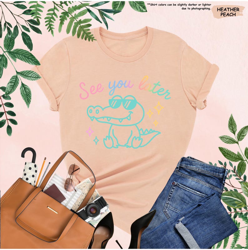 May include: A peach-colored t-shirt with the text "See you later" and a cartoon alligator wearing sunglasses. The shirt is paired with blue jeans, a brown tote bag, black heels, and accessories. Green foliage surrounds the items.