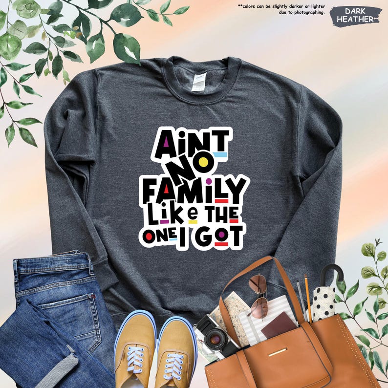May include: Dark heather gray sweatshirt with the text "Ain't No Family Like The One I Got" in a colorful, bold font. The image includes blue jeans, tan shoes, a brown tote bag, and accessories.