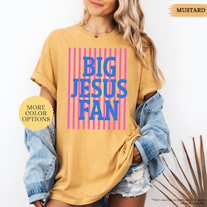 May include: Mustard-colored t-shirt with the text "BIG JESUS FAN" in blue letters, set against a pink striped background. The shirt has rolled-up sleeves and is part of a clothing collection.