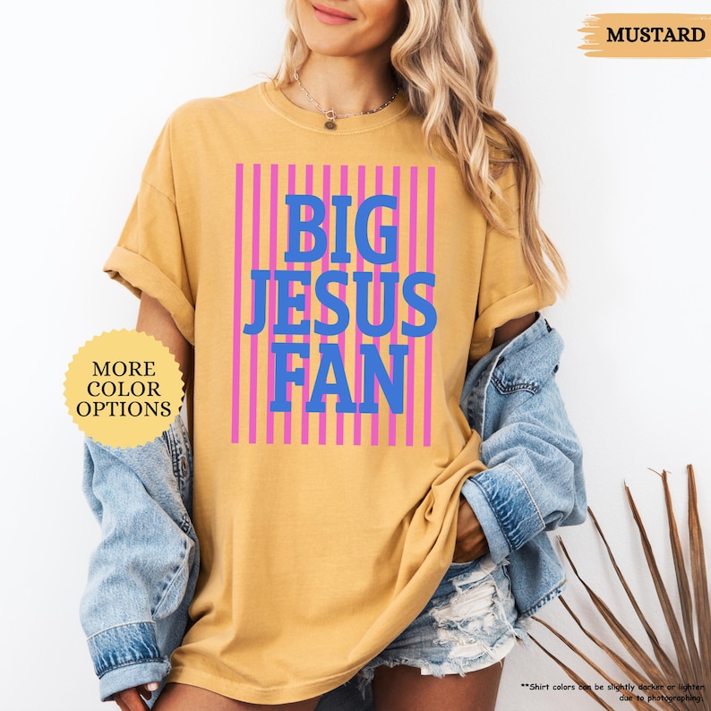 May include: Mustard-colored t-shirt with the text "BIG JESUS FAN" in blue letters, set against a pink striped background. The shirt has rolled-up sleeves and is part of a clothing collection.