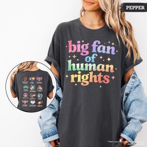 May include: Dark gray t-shirt with the text "big fan of human rights" in colorful letters. The back of the shirt features various icons and phrases related to human rights. The shirt is a comfortable, casual style.