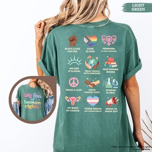 May include: Light green t-shirt with various graphic designs and text. The shirt features phrases like "Black Lives Matter," "Love is Love," and "Speak Up, Speak Out." A smaller image on the shirt says "big fan of human rights."