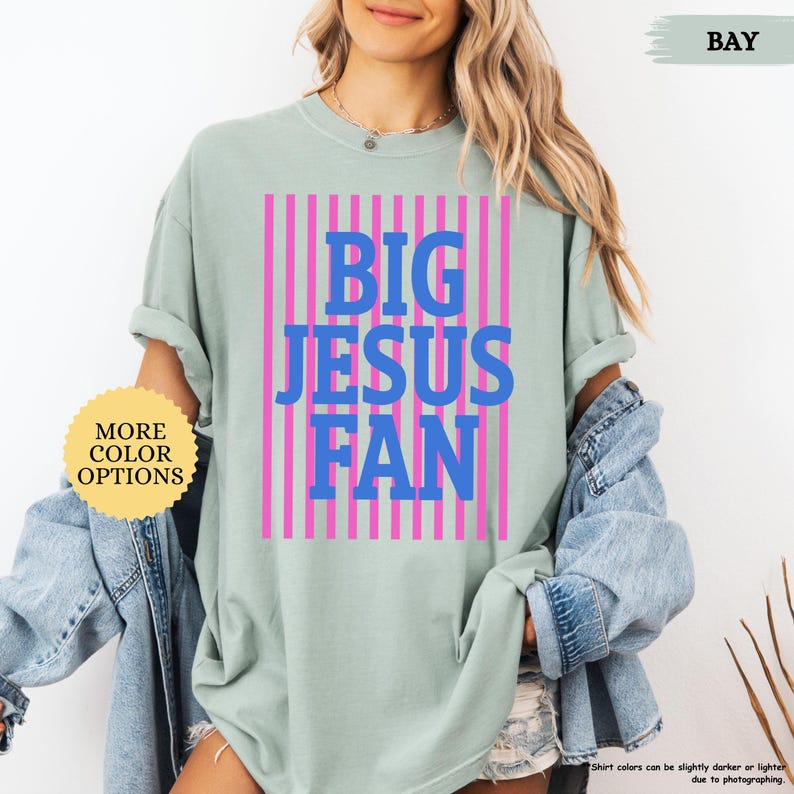May include: A light green t-shirt with the text "BIG JESUS FAN" in blue letters, set against a pink striped background. The shirt also has a "MORE COLOR OPTIONS" button.